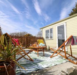 An outdoor patio area with wooden deck chairs around a small wooden table on a patterned rug next to a light green mobile home at Cwrt y Ceffyl Boduan near Morfa Nefyn