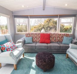 A living room with a gray sofa and two light blue armchairs with colorful pillows and a round dark ottoman on a light blue carpet at Cwrt y Ceffyl in Boduan near Morfa Nefyn