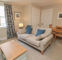 A living room with a beige sofa with blue cushions a wooden coffee table curtains on the window and a small dining table with chairs at Charter's Cottage in Berwick-Upon-Tweed