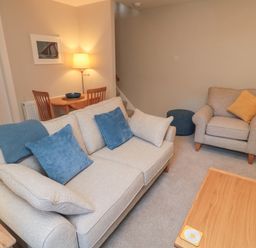 A living room with beige sofa with blue cushions armchair with yellow cushion coffee table and small dining table with lamp at Charter's Cottage in Berwick-Upon-Tweed
