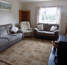 A living room with two sofas, a beige armchair, a wooden corner shelf, a window with curtains, and a fireplace with a stove at Brookfield in St Gennys near Wainhouse Corner