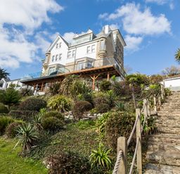 A stone stairway with rope railing leading up to a large house on a hill with plants and shrubs at The Tower 5 Corisande Manor in Newquay