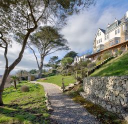 A gravel path next to stone retaining walls with trees grass and a multi story building on a hill at The Tower 5 Corisande Manor in Newquay