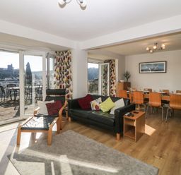 A living room with a black sofa and wooden chair next to large glass doors leading to a balcony and a dining area with a wooden table and chairs in Estuary View in Newton Ferrers