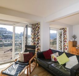 A living room with black leather sofa and chair with cushions a wooden dining table and chairs near large glass doors with a balcony and hillside view at Estuary View in Newton Ferrers