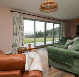 A living room with brown and green sofas a wooden side table with books and a plant large glass sliding doors with floral curtains showing a grassy outdoor area at Dol Blodau in Newbridge-On-Wye