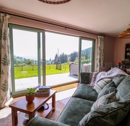 A living room with a green sofa, wooden coffee table, and large sliding glass doors showing an outdoor patio and field at Dol Blodau in Newbridge-On-Wye