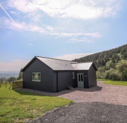 A single-story dark wooden house with a gravel driveway surrounded by grass and trees at Ystrad Wen in Llysdinam near Newbridge-On-Wye