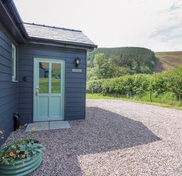 The exterior of a building with a light blue door a black wall light and a flower planter on a gravel driveway with trees and hills in the background at Ystrad Wen in Llysdinam near Newbridge-On-Wye