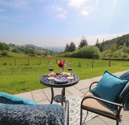 A patio with two chairs and a table set with wine glasses and food overlooking a grassy field and hills at Ystrad Wen in Llysdinam near Newbridge-On-Wye