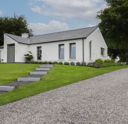 A single-story white house with a gray roof and three windows on a grassy lawn with stone steps and a gravel driveway at Shannon Vale in Woodford County Galway