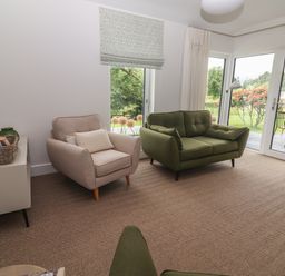 A sitting room with a beige armchair and a green loveseat near glass doors overlooking a garden at Shannon Vale in Woodford County Galway