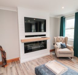 A living room with a mounted TV above a fireplace an armchair with cushions green curtains and a tray on an ottoman at 2 Apple Tree in Beadnell