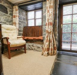 A corner of a room with a beige padded chair wooden bench under windows and a glass door with floral curtains at The Searle in Nantmel near Rhayader
