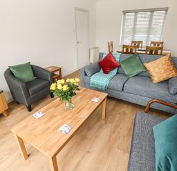 A living room with a wooden coffee table yellow flowers on the table sofas and armchairs with cushions and a dining table with chairs at Timaru in Lamphey