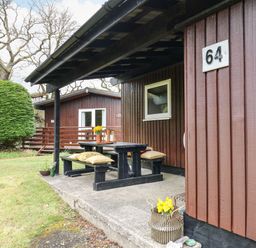 A covered outdoor seating area with a wooden picnic table and benches with cushions and yellow flowers at 64 Penlan Holiday Park in Cenarth