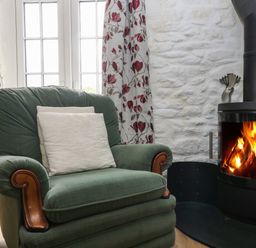 A green armchair with white cushions near a wood burning stove and floral curtains at Byre Trethin near Camelford
