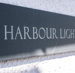 A black sign with white letters reading Harbour Lights mounted on a white textured wall at Harbour Lights Port Isaac