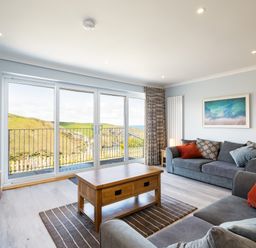 A living room with two grey sofas cushions a wooden coffee table carpet tv on a wooden stand and sliding glass doors with a view at Harbour Lights Port Isaac