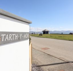 A road with a small pavilion and people sitting on benches near a grassy area by the sea at Tarth Y Mor in Criccieth
