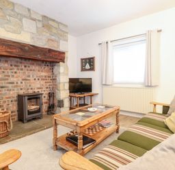 A living room with a stone and brick fireplace a wood stove a wooden coffee table and a striped sofa at Canny Cottage Low Hauxley Nr Amble