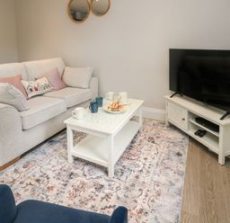 A living room with a white sofa pink and patterned cushions a white coffee table with mugs and pastries a flat screen tv on a white tv stand and a patterned rug at 3 Compass Point in Weymouth
