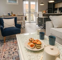 A living area with a beige sofa and blue armchair around a white coffee table with muffins and cups in a kitchen dining space at 3 Compass Point in Weymouth