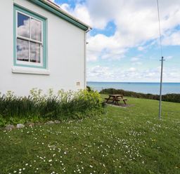 Side of a white house with a window and garden next to a lawn with a picnic table and a sea view at Beachcliff in Gorran Haven