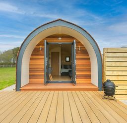 A wooden pod structure with open double doors leading to an interior with a bed and chairs on a deck at Sun Rise in St Buryan