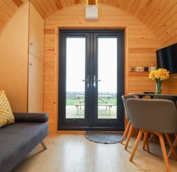 A small wooden room with a grey sofa and yellow cushion beside a cabinet and glass doors with a table and chairs on the right with a vase of flowers and a TV mounted on the wall at Sun Rise in St Buryan