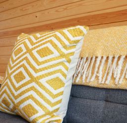 A yellow and white patterned cushion and a yellow and white fringed blanket on a grey sofa with a wooden wall behind at Sun Rise in St Buryan