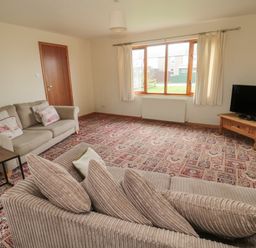 A living room with beige sofas a patterned carpet a wooden door a window with curtains and a TV on a wooden stand at Causeway Cottage in Holy Island