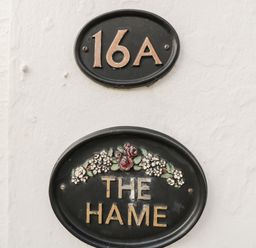 Black oval house number plaque with 16A above a black oval plaque with The Hame and floral decoration on a white wall at The Hame in Monmouth