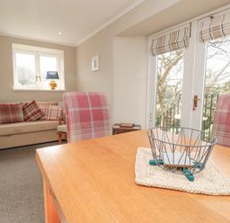 A dining table with a wire basket and chairs beside a sofa with plaid cushions in a living room with windows and a door at Magnolia House in Dittisham