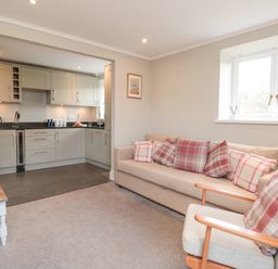 A living room with a beige sofa and plaid cushions next to a window with a lamp and an armchair adjacent to a kitchen with cabinets and appliances at Magnolia House in Dittisham
