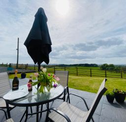 An outdoor patio with a glass table and chairs a bouquet of flowers and bottles on the table a lawn and wooden fence with a view of fields and trees at Dryfe Lodge in Lockerbie