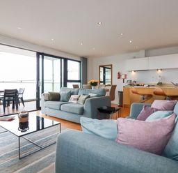 A living room with blue sofas and pink cushions a coffee table and a dining area with kitchen in the background at Fistral Pearl in Newquay