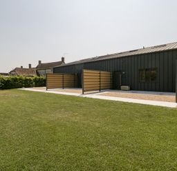 A modern dark-colored building with fenced patio areas and a lawn in front at Linden Barn in Wincanton