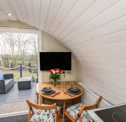 A small dining area with a table set for two and a TV on the wall next to an open door leading to an outdoor seating area at Cherry Pod 2 Hookagate near Meole Brace