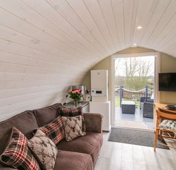 A room with a brown sofa with cushions a small dining table with two chairs a wall-mounted TV and a door leading outside at Cherry Pod 2 Hookagate near Meole Brace