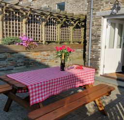 A wooden picnic table with a red and white checkered tablecloth with flowers and wine outside a stone building with open door at The Mill in Trethevy near Tintagel