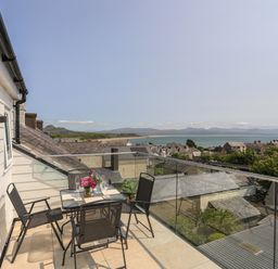 A balcony with a table and four chairs with flowers and glasses overlooking a town and the sea at Golygfa'r Gastell Castle View in Criccieth