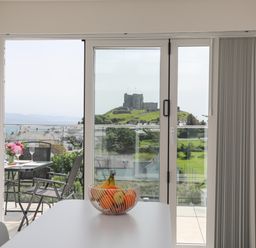 A dining area with a bowl of fruit on the table and a balcony with chairs and flowers overlooking a castle at Golygfa'r Gastell Castle View in Criccieth