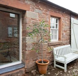 A stone wall with windows a door a potted plant and a white bench at The Courtyard in Kirklinton near Longtown
