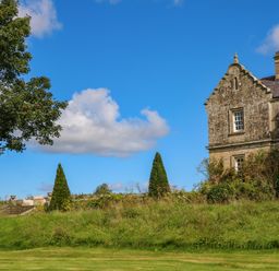 A stone building with a lawn trees and bushes under a blue sky at The Pavillion in Kirklinton near Longtown