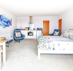 A bedroom with a double bed floral bedding two blue chairs a small kitchen unit and a dining table at Greenside Studio in Praa Sands