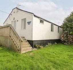 A white mobile home with steps leading to a door on a grassy lawn with bushes at Suzannes Beach Hut in Bacton near Walcott