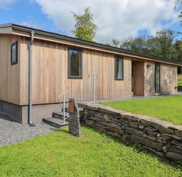 A single-story wooden house with black-framed windows a small staircase and a stone wall in a grassy yard at Millstream Ridge in Haverthwaite