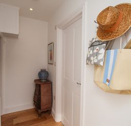 A hallway with a chest of drawers and a blue vase a closed white door and hats and bags hanging on wall hooks at The Corner House in Weymouth