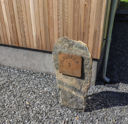 A stone with a metal plaque showing The Burrow 2 outside a building with wooden siding and gravel ground at The Burrow in Haverthwaite